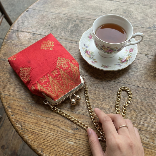 Red handbag with gold chain on a wooden table next to a cup of tea.